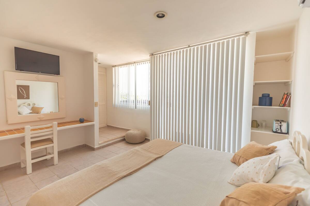 A bedroom with a bed, a desk, a chair, a mirror, and a window with blinds.
