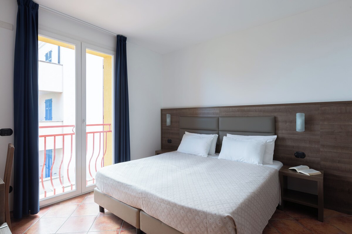 A hotel room with a large bed, a wooden headboard, a nightstand with a phone and book, and a balcony with red railings.