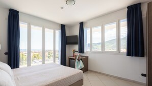 A hotel room with a large bed, a TV mounted on the wall, a small desk with a plant, and a view of the city and hills through large windows. - Ai Pozzi Village Resort & SPA (Loano)