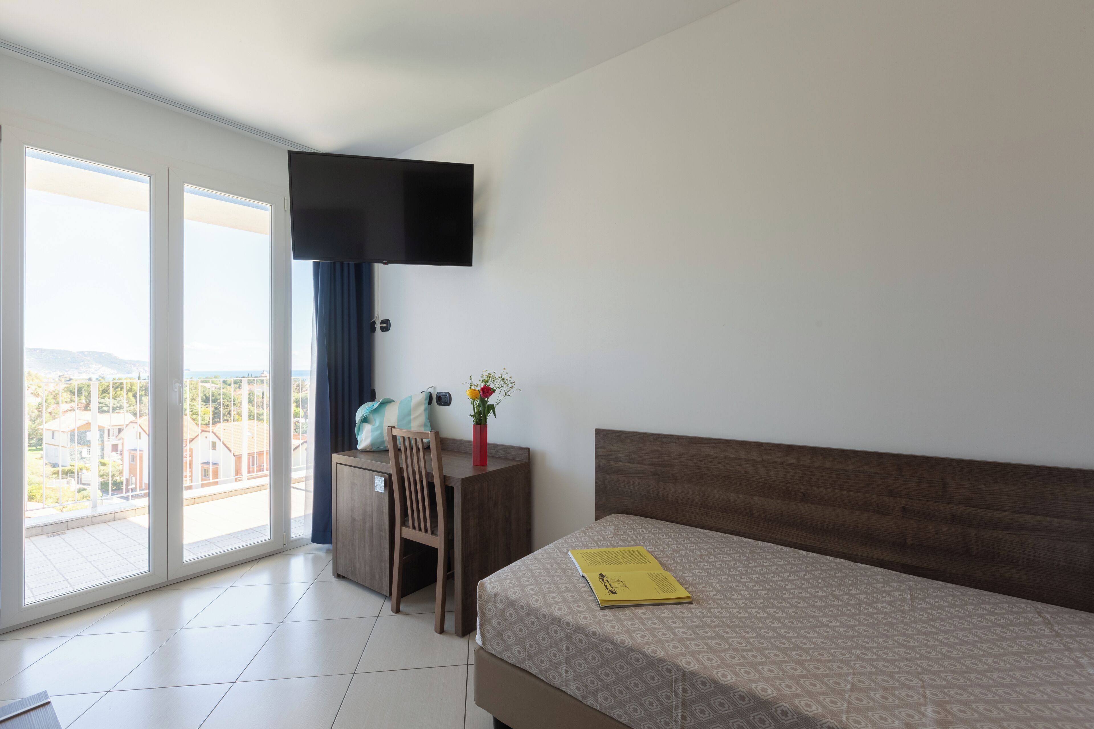 Habitación de hotel con cama, escritorio, silla, televisor de pantalla plana montado en la pared y un amplio ventanal con vistas a edificios y una montaña.