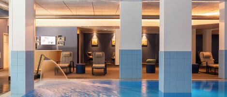 An indoor swimming pool with a blue and white tiled floor, surrounded by lounge chairs and a wall with framed artwork.