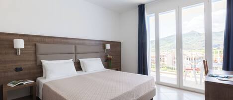 A hotel room with a large bed, a desk, and a view of the city through large windows.