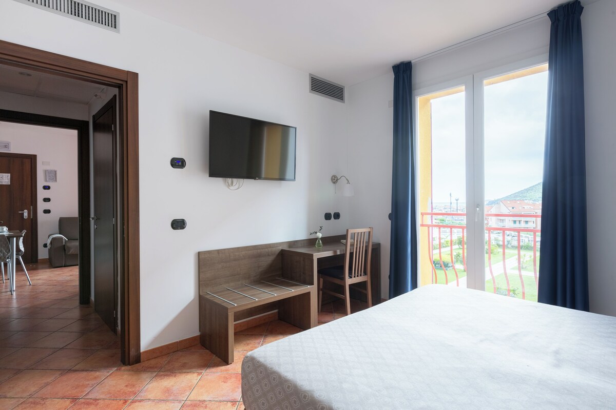 A hotel room with a bed, a desk, a chair, a TV, and a balcony with a view.
