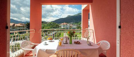 Un balcon avec une table dressée pour le petit-déjeuner, des chaises blanches et une vue sur les bâtiments et les montagnes environnants.