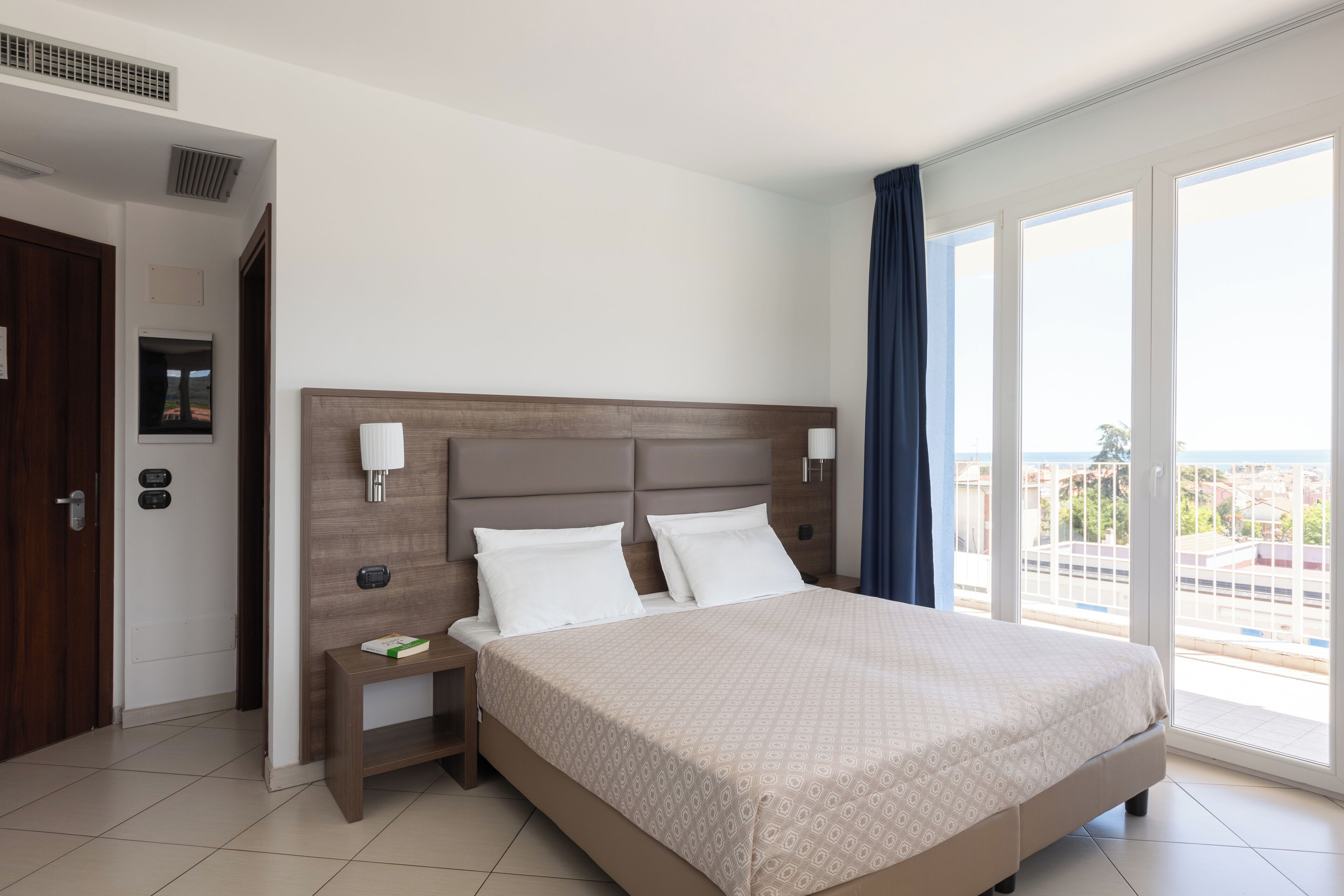Una habitación de hotel con una cama grande, una mesita de noche, una lámpara de pared, un ventanal con vistas y una puerta con picaporte.