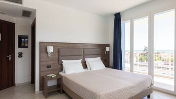 Une chambre d’hôtel avec un grand lit, une table de chevet, une lampe fixée au mur, une fenêtre donnant sur un beau paysage et une porte avec une poignée.