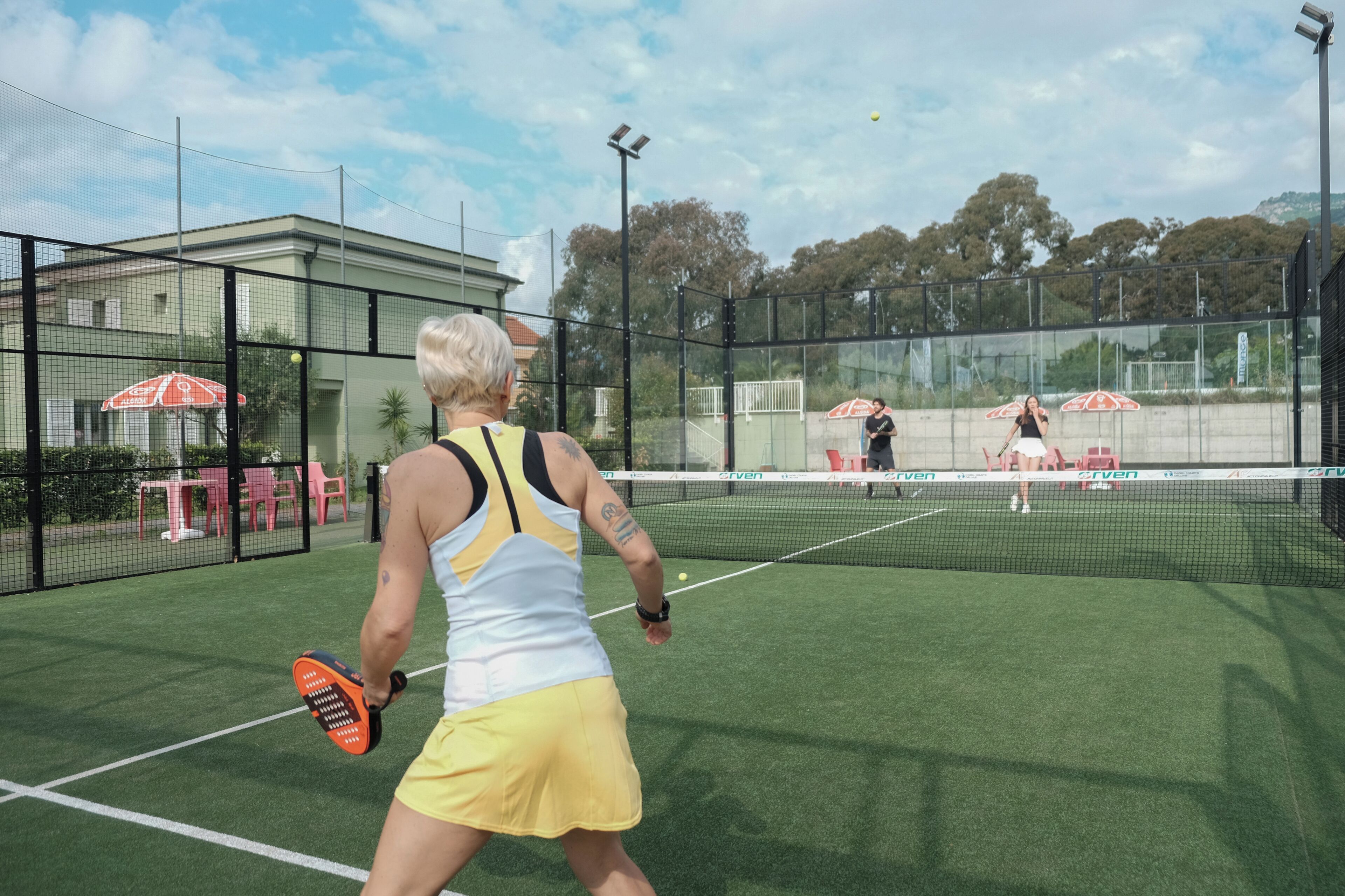 Un court de tennis avec un filet, un joueur vêtu d’une tenue jaune et un bâtiment en arrière-plan.