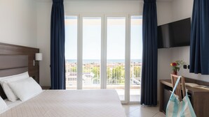 A hotel room with a large bed, a desk with a chair, a TV mounted on the wall, and a view of the city through the windows. - Ai Pozzi Village Resort & SPA (Loano)