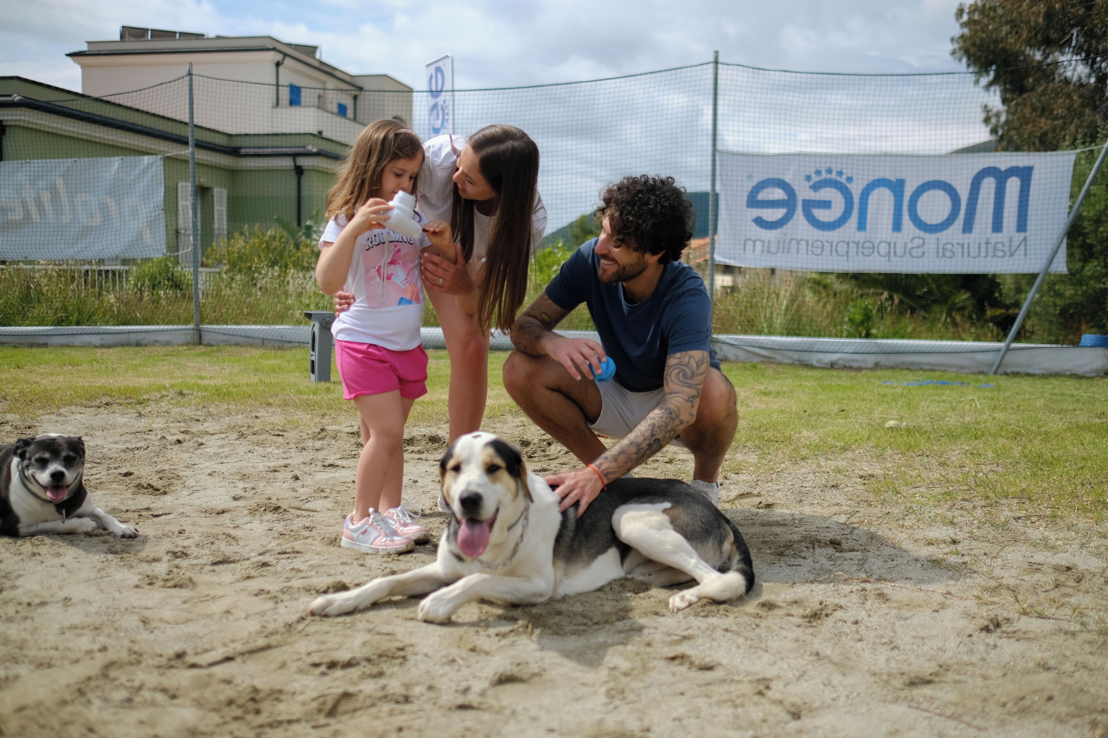 A sandy outdoor area with a dog lying down, a person crouching, and another person standing.