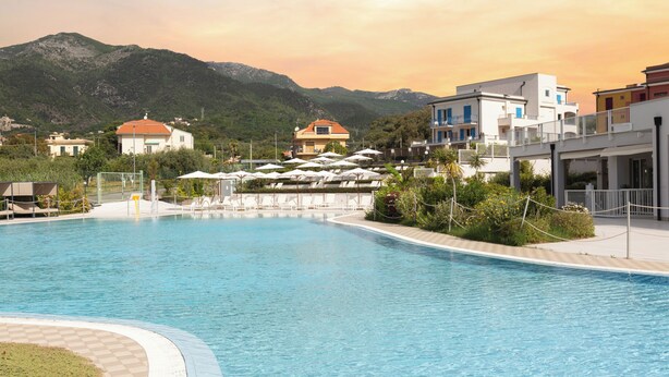 A swimming pool with lounge chairs and umbrellas, surrounded by buildings and mountains. - Ai Pozzi Village Resort & SPA (Loano)