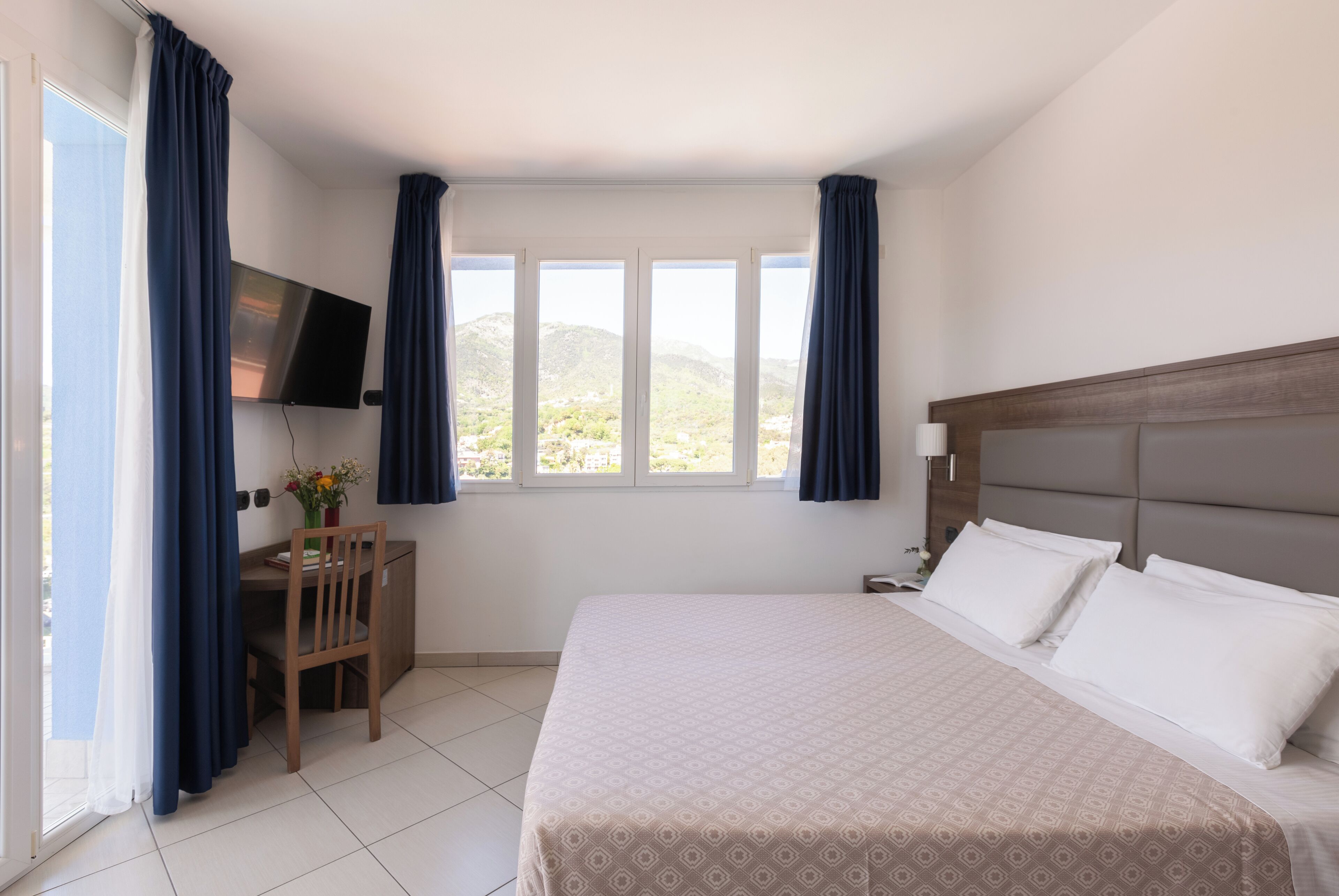 A hotel room with a bed, a desk, a chair, a TV, and a view of the mountains.