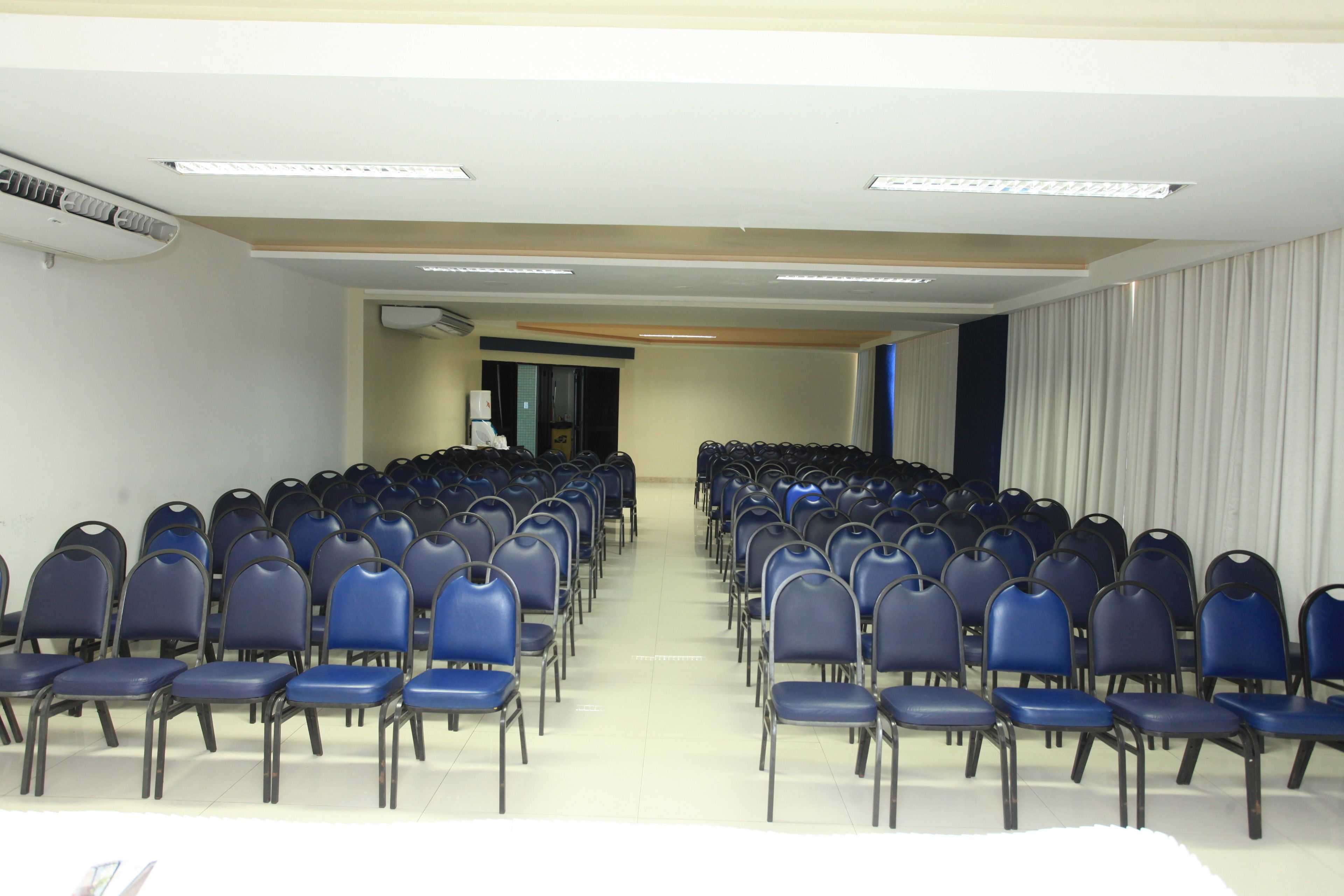 Un salón de conferencias con filas de sillas azules, una mesa blanca y una pantalla de proyector al frente.