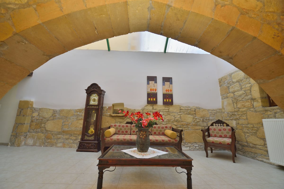 A cozy room with a stone wall, a wooden sofa, a wooden chair, a table with a vase of flowers, and a grandfather clock.