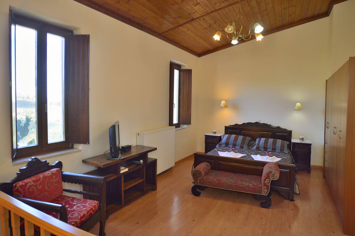 A bedroom with a wooden ceiling, a bed with patterned bedding, a desk with a computer, a chair, and a window with blinds.