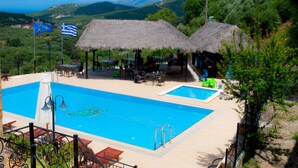 A pool area with two thatched-roof structures, a view of the landscape, and a flag. - Maria Villas (Kissamos)