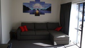 A living room with a sectional sofa, a multi-panel artwork, and a view through a window.