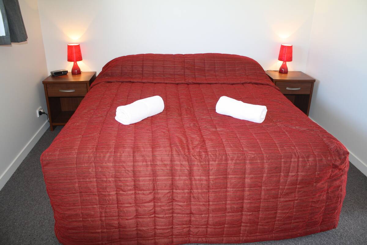 Ein ordentlich bezogenes Bett mit einer roten Decke, zwei Nachttische mit Lampen und eine Steckdose an der Wand.