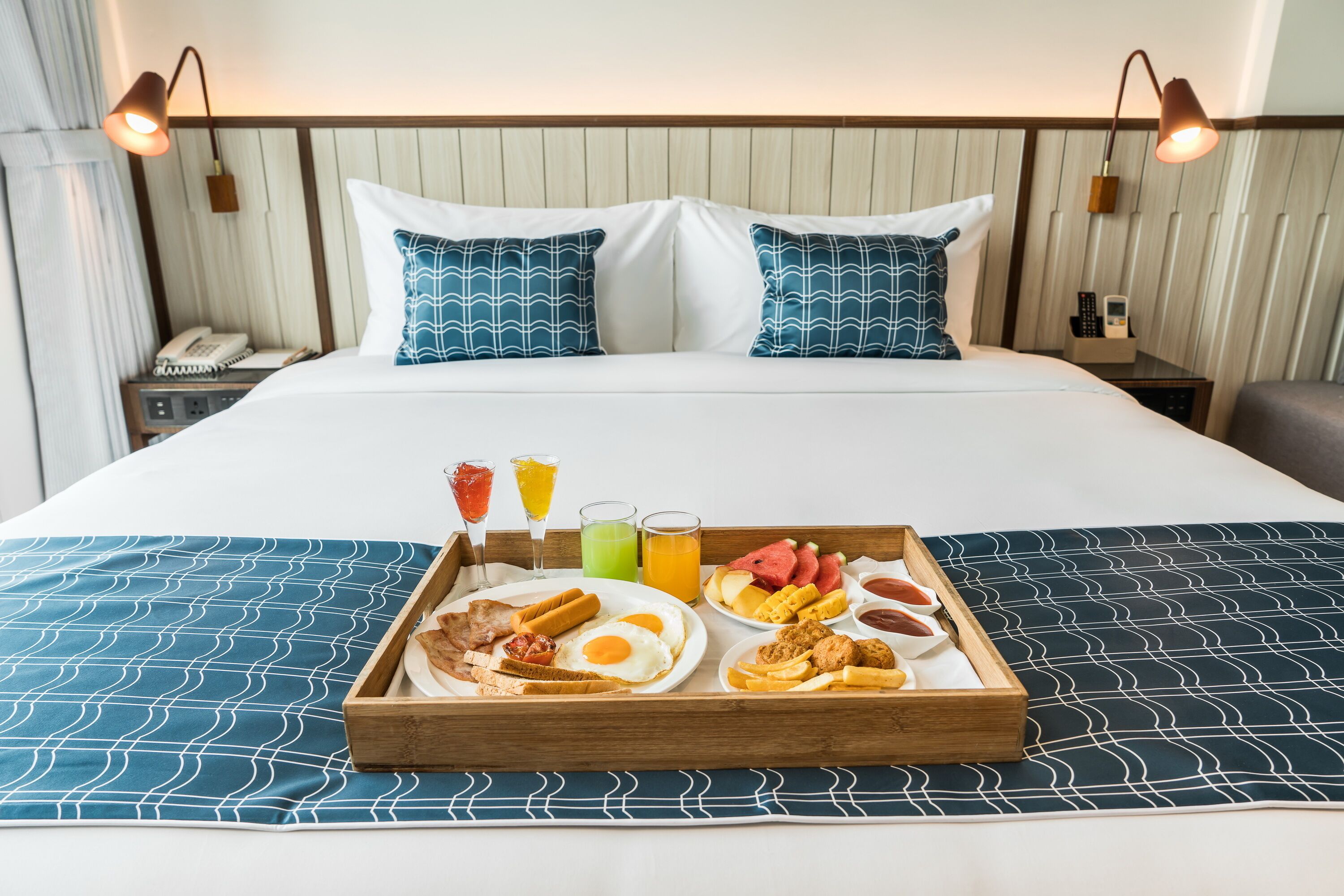 A hotel room with a neatly made bed, a tray of breakfast including eggs, bacon, sausages, and fruit, and a bedside table with a phone and a lamp.