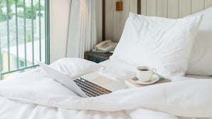 Una cama bien hecha con sábanas blancas, una computadora portátil, una taza de café y una mesita de noche con un teléfono.