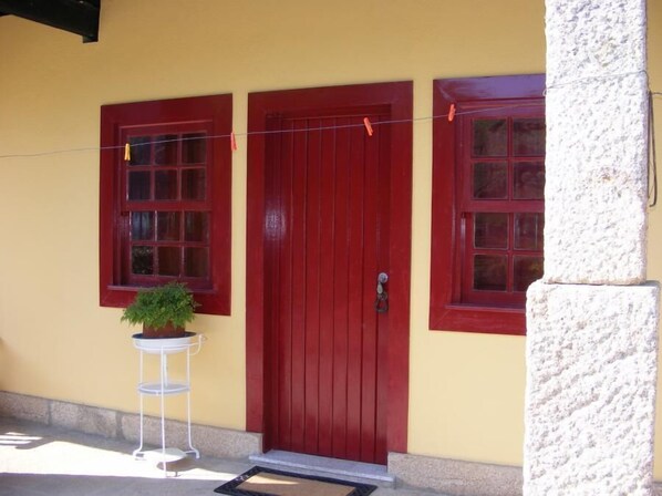 A building with a red door and two windows, a small white table with a plant, and a doormat. - Solar Quinta da Portela (Santa Marta de Penaguião)