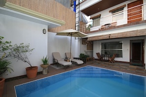Una piscina con sillones de descanso y una mesa, rodeada de plantas en macetas y una terraza de madera.