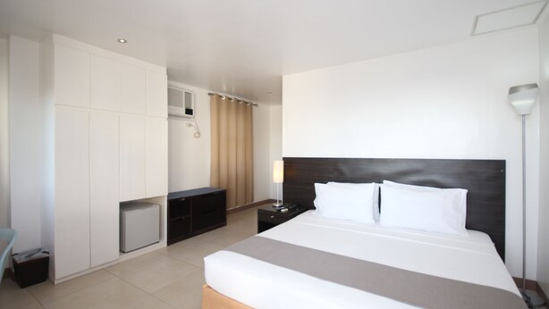 A modern hotel room with a large bed, a desk, a wardrobe, and a small refrigerator.