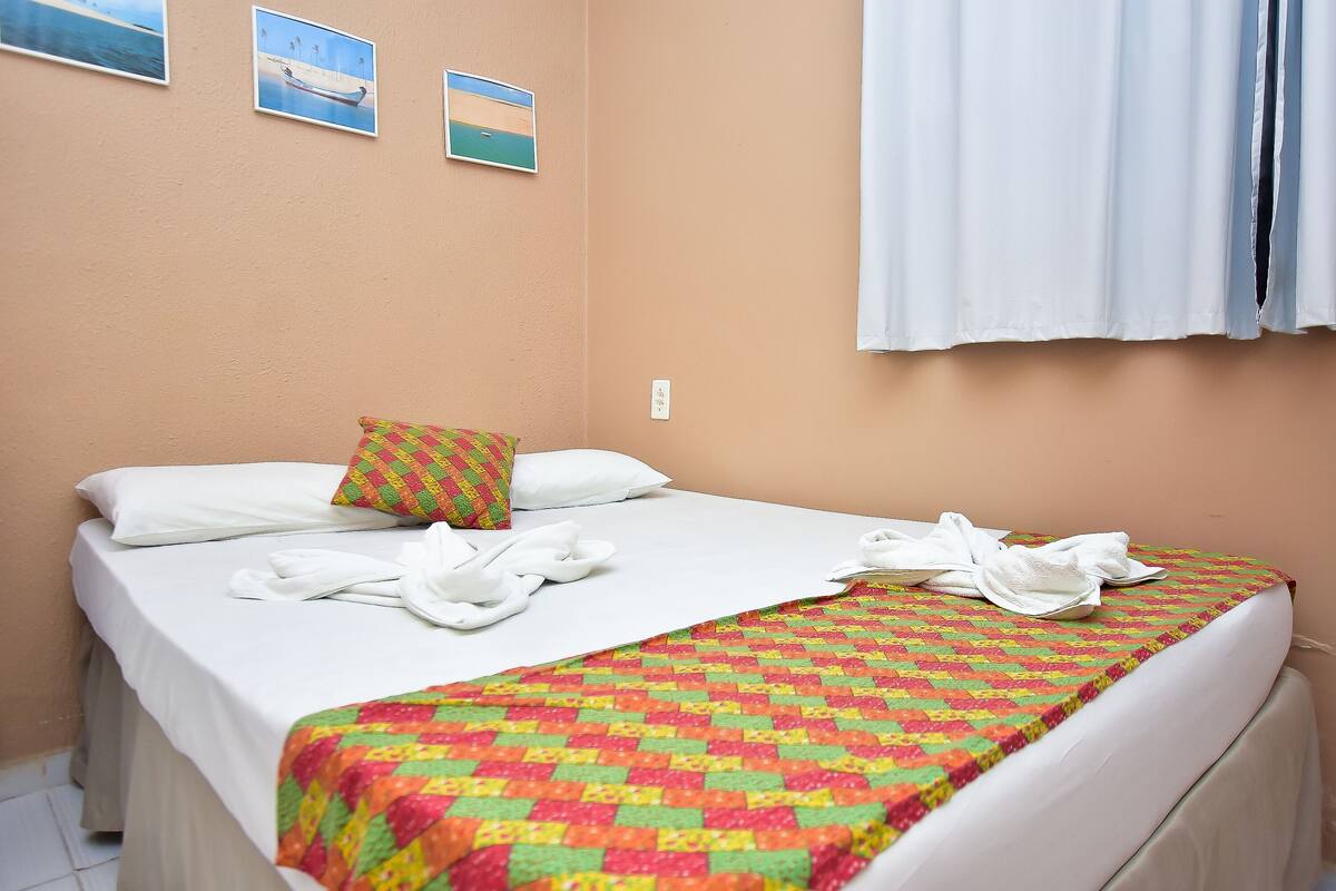 A neatly made bed with white linens, a colorful quilt, and a white towel. There are two pillows, one plain white and one with a checkered pattern. The room has peach-colored walls and a window with white curtains.