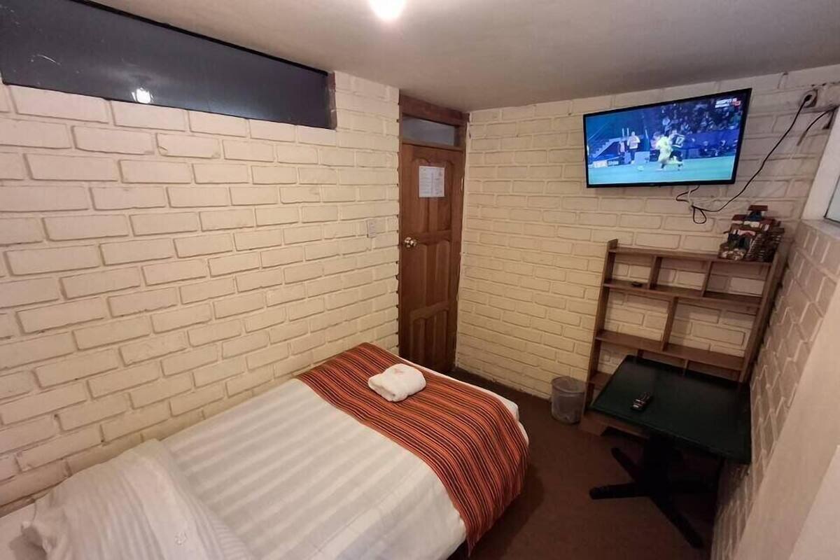 A small, compact room with a single bed, a TV mounted on the wall, and a small desk with a chair.