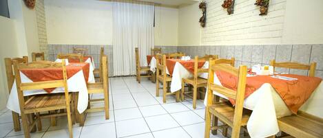 A dining area with wooden tables and chairs, white tiled floor, and a brick wall with flower baskets.