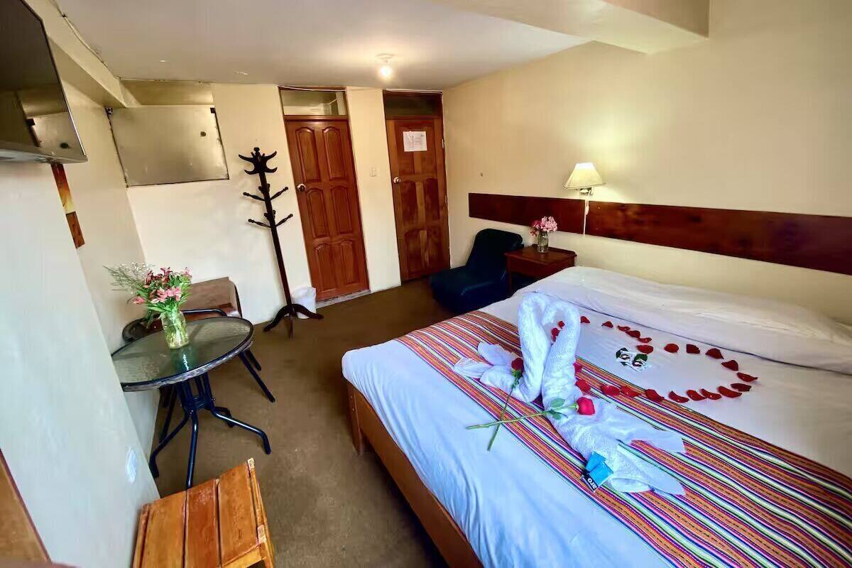 A hotel room with a bed, a desk with a chair, a small table with a vase of flowers, and a door.