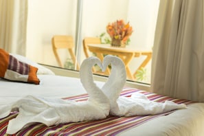 A bed with a towel arranged in the shape of a swan, a striped bedspread, and a window with a view of a dining area. - Andean Dreams Hotel (Cusco)