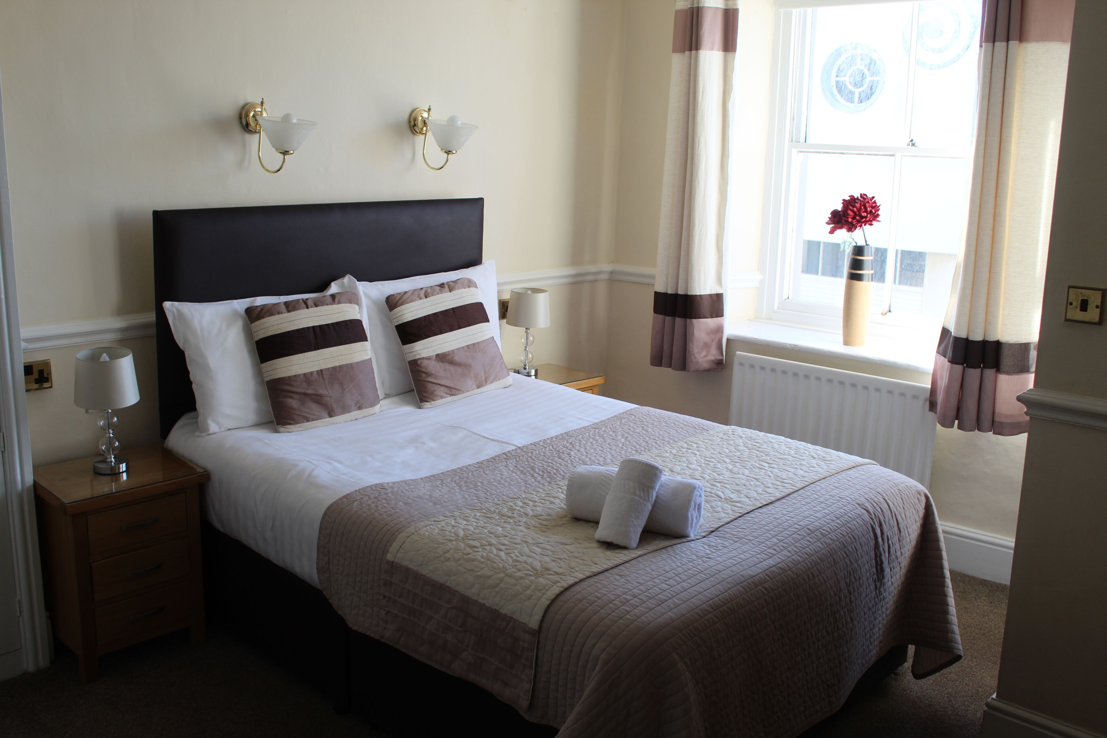 A neatly made bed with a headboard, two bedside lamps, a nightstand, and a window with curtains.