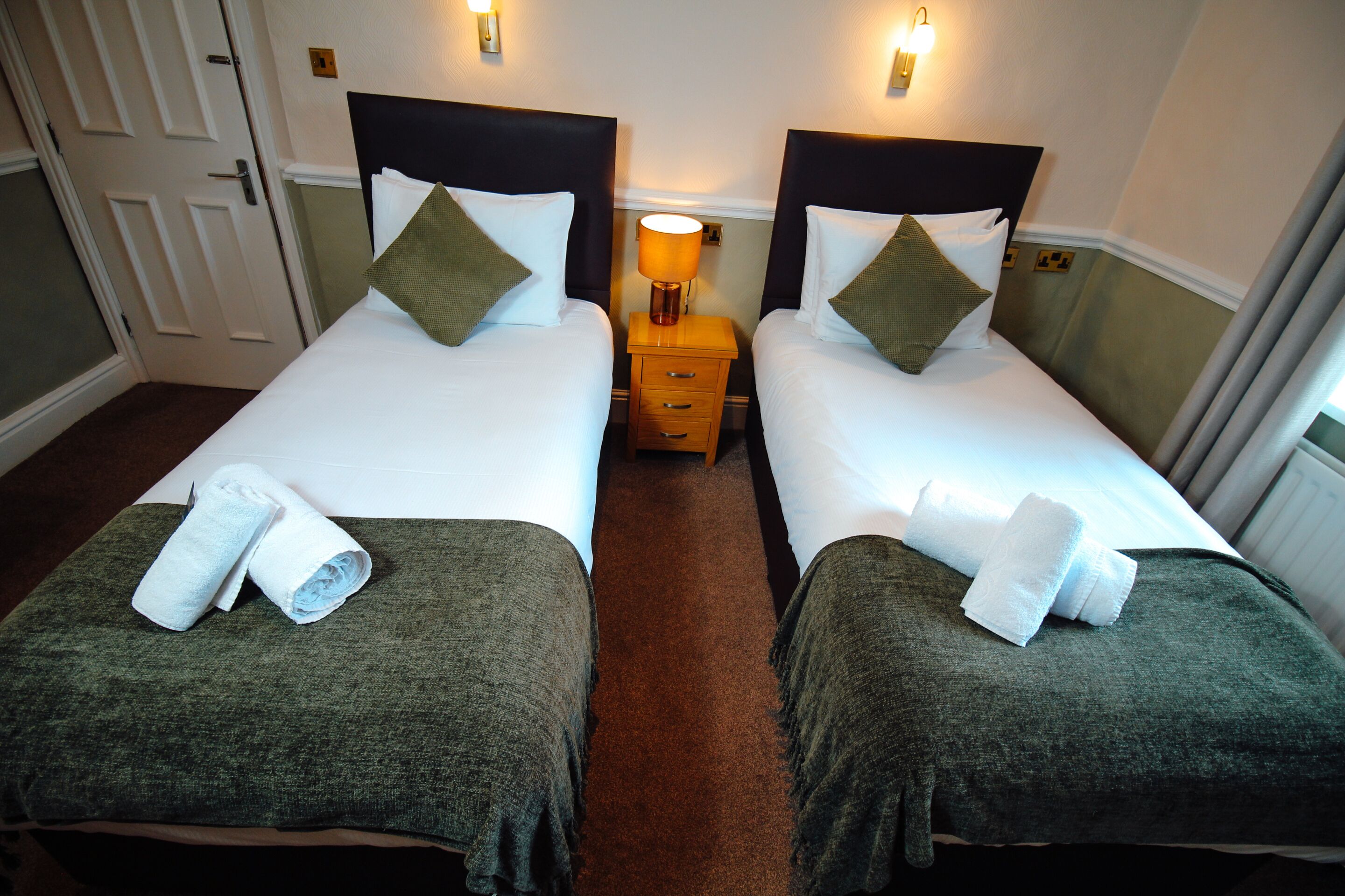 A hotel room with two beds, a nightstand with a lamp, and a door.