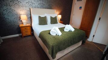 Una cama bien hecha con una colcha verde, dos almohadas y toallas enrolladas. Se ve un mesita de noche con una lámpara y un cajón de madera. La habitación tiene papel tapiz estampado y una puerta de madera.