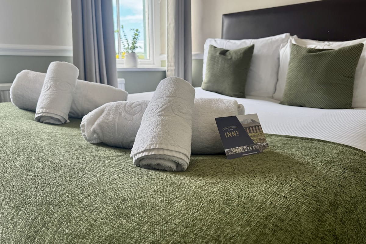 A neatly made bed with white linens and a green throw blanket, a pillow, and a tag indicating the hotel name.