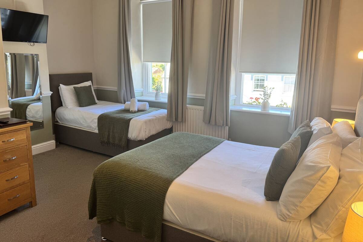 A hotel room with two beds, a TV, a dresser, and large windows with curtains.