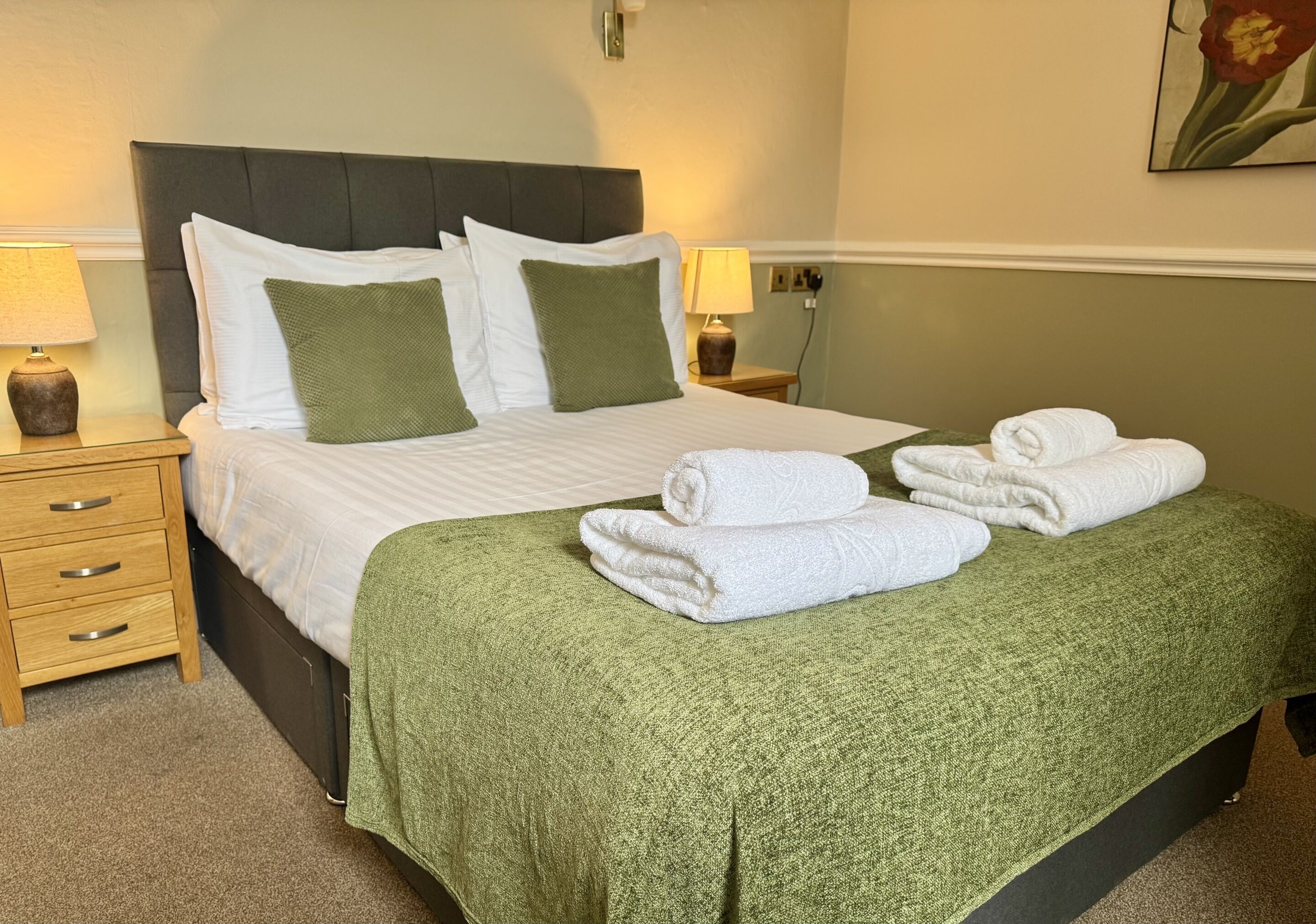 A neatly made bed with white linens and green pillows, a green throw blanket, and folded towels on the foot of the bed.