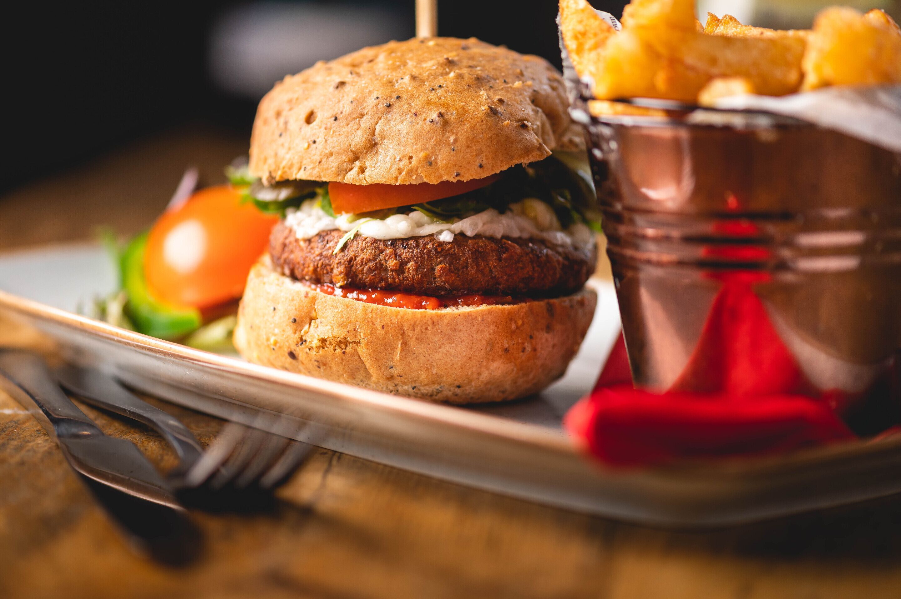 A burger with a bun, lettuce, tomato, and a patty, served with fries and ketchup in a copper cup.