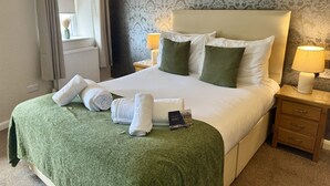 Ein ordentlich bezogenes Bett mit einer grünen Decke, weißen Kissen und zusammengerollten Handtüchern. Ein Nachttisch mit einer Lampe und einem Buch mit dem Titel 