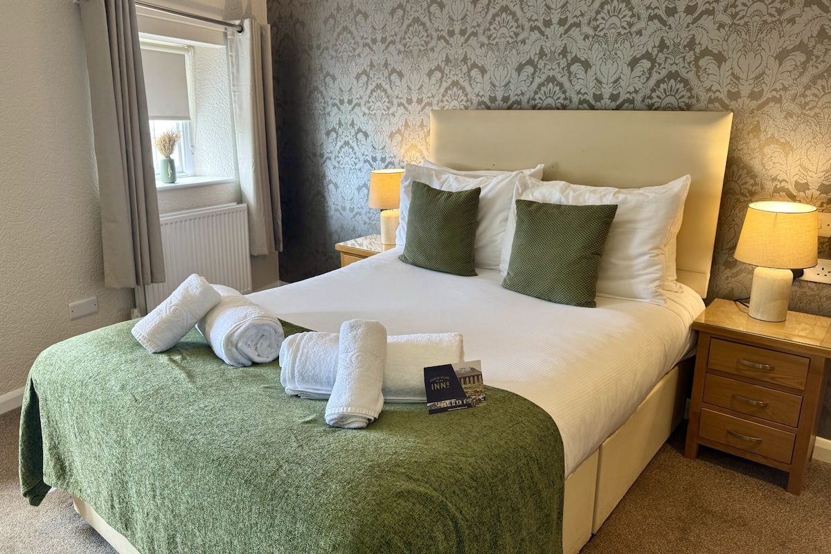 A neatly made bed with a green blanket, white pillows, and rolled towels. There is a nightstand with a lamp and a book titled "Inn Hero".