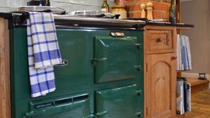 A green vintage stove with a checkered towel hanging on it, a brick wall, and a wooden cabinet with books. - Motts Cottage (Dunmow)