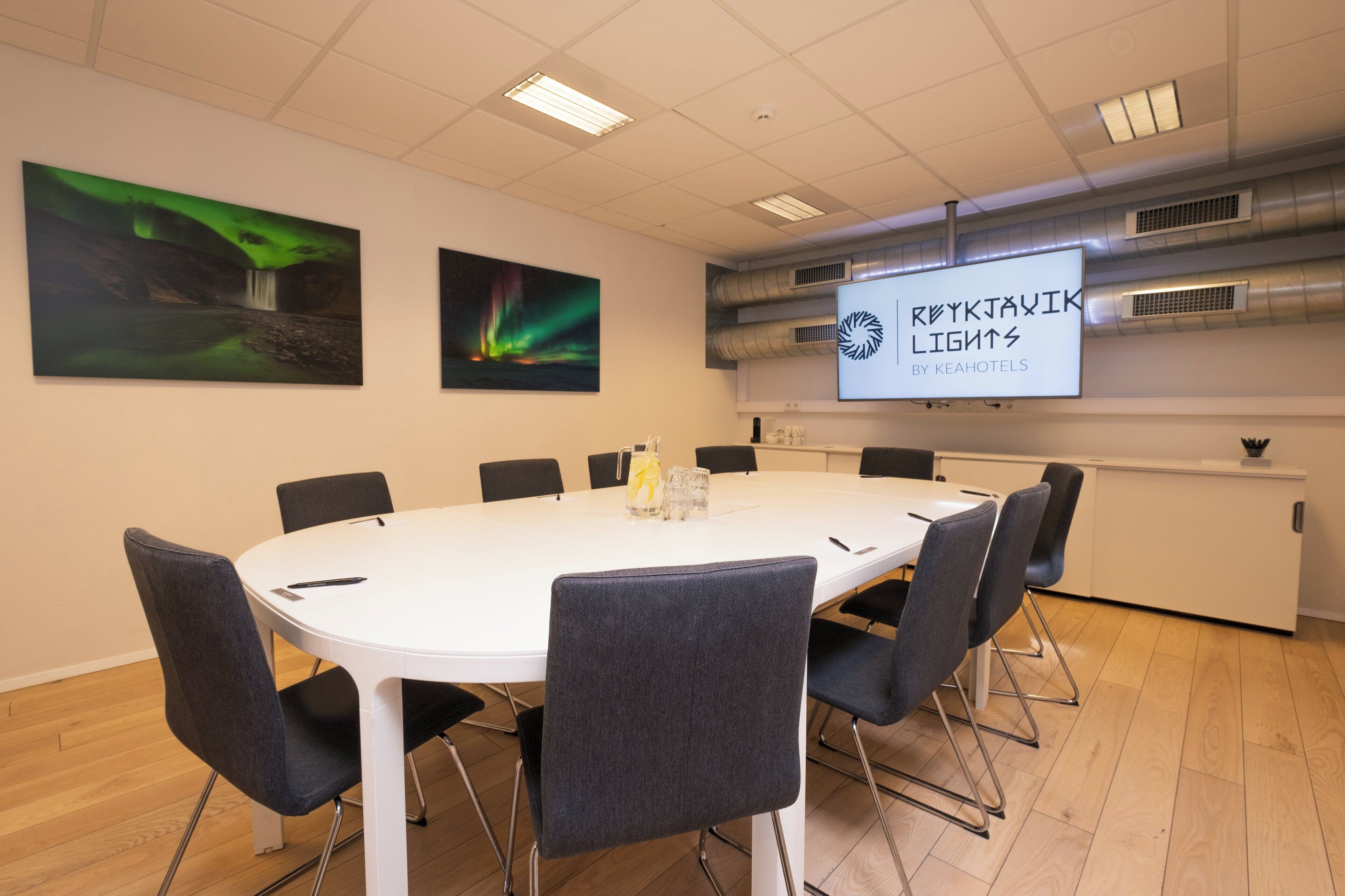 A conference room with a white table, grey chairs, and a projector screen displaying 'RØYKJAKJIK LIGHTS BY KEAHOTELS'.
