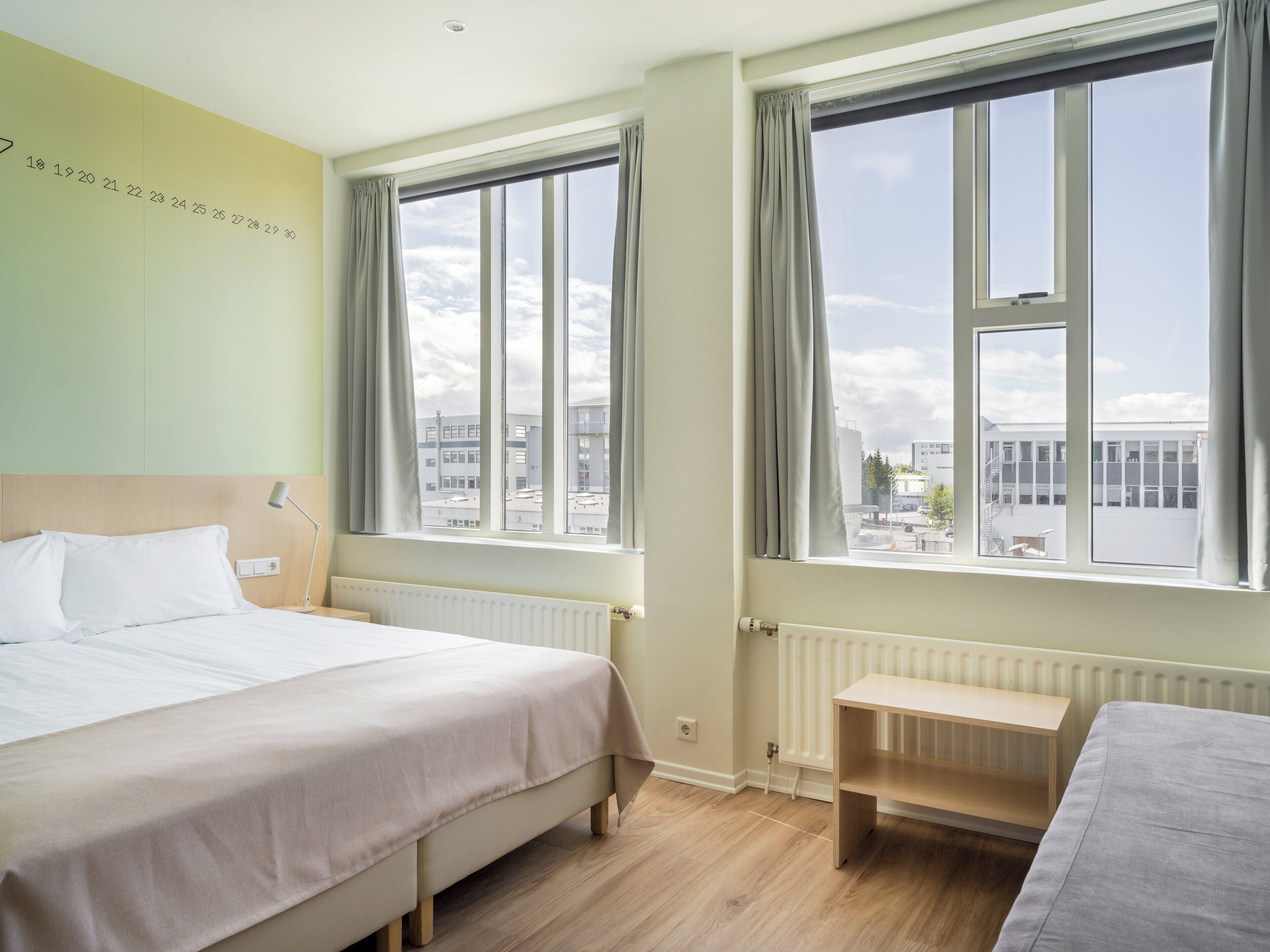 A hotel room with a bed, a bedside table, a bench, and large windows offering a city view.