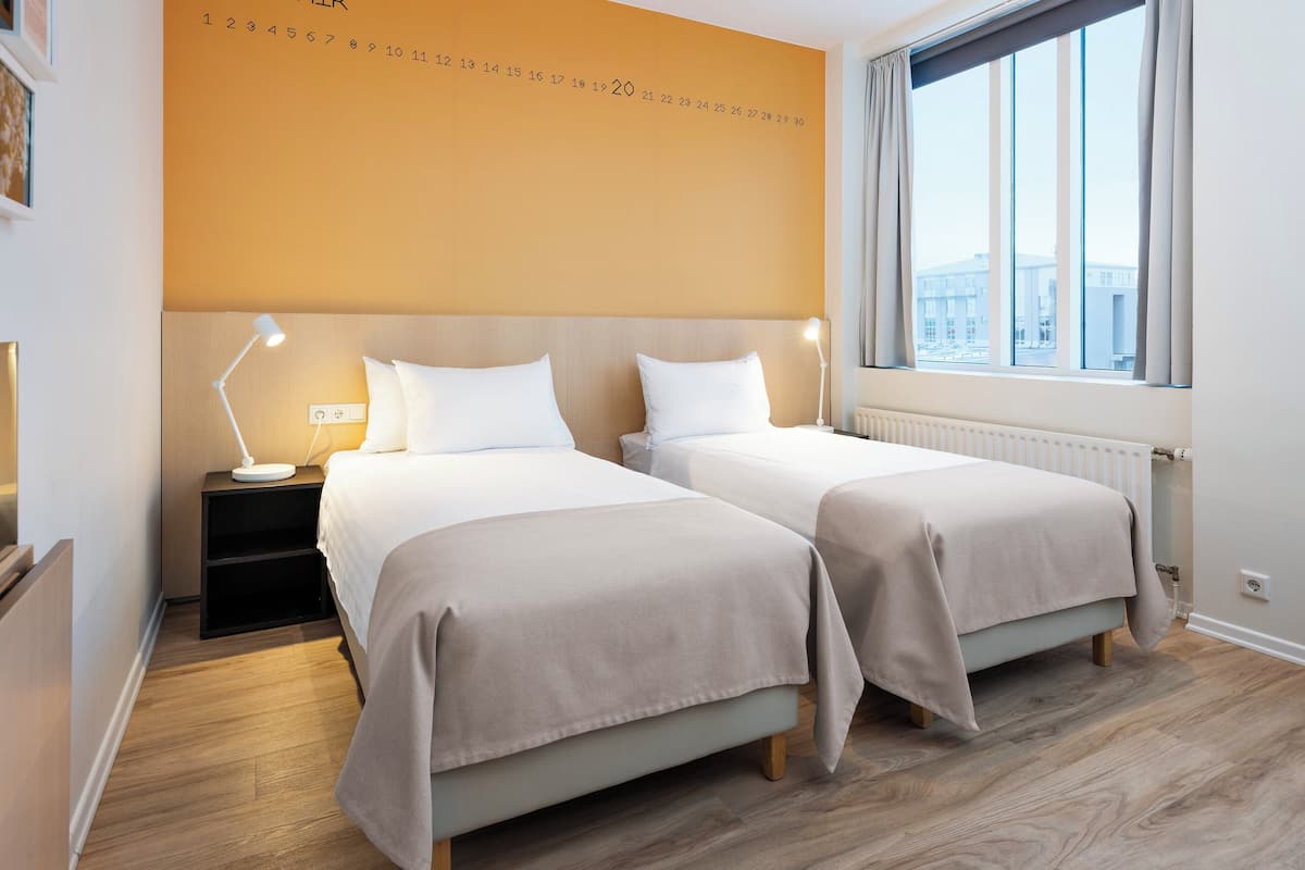 A hotel room with two beds, a nightstand, a desk lamp, a window with a view of buildings, and a wall calendar.