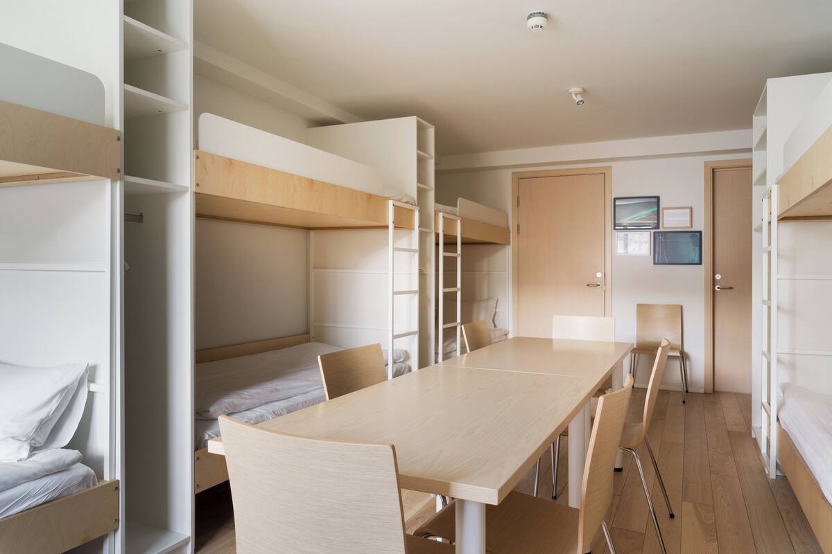 A dormitory room with bunk beds, a desk, chairs, and a door.