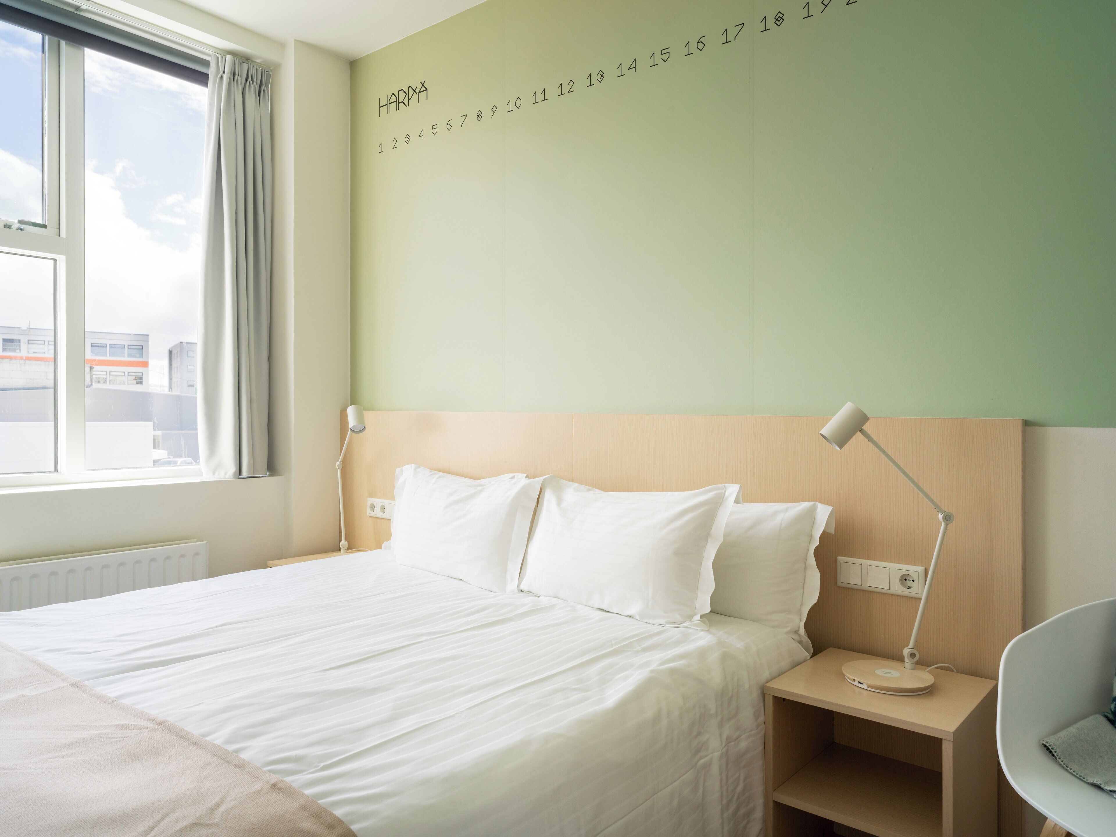 A hotel room with a large bed, a nightstand, a desk lamp, and a view of the city through the window.