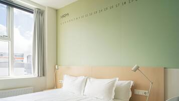 A hotel room with a large bed, a nightstand, a desk lamp, and a view of the city through the window.