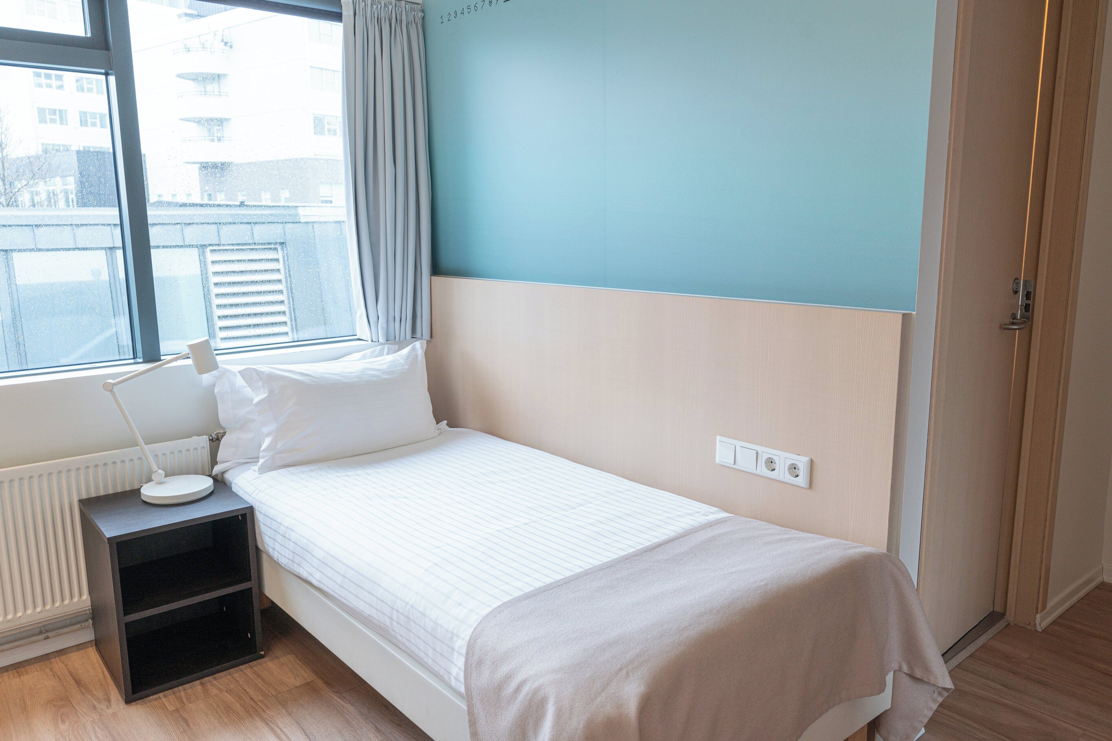 A single bed with a white comforter and pillows, a bedside table with a lamp, a power outlet, and a window with a view of buildings.