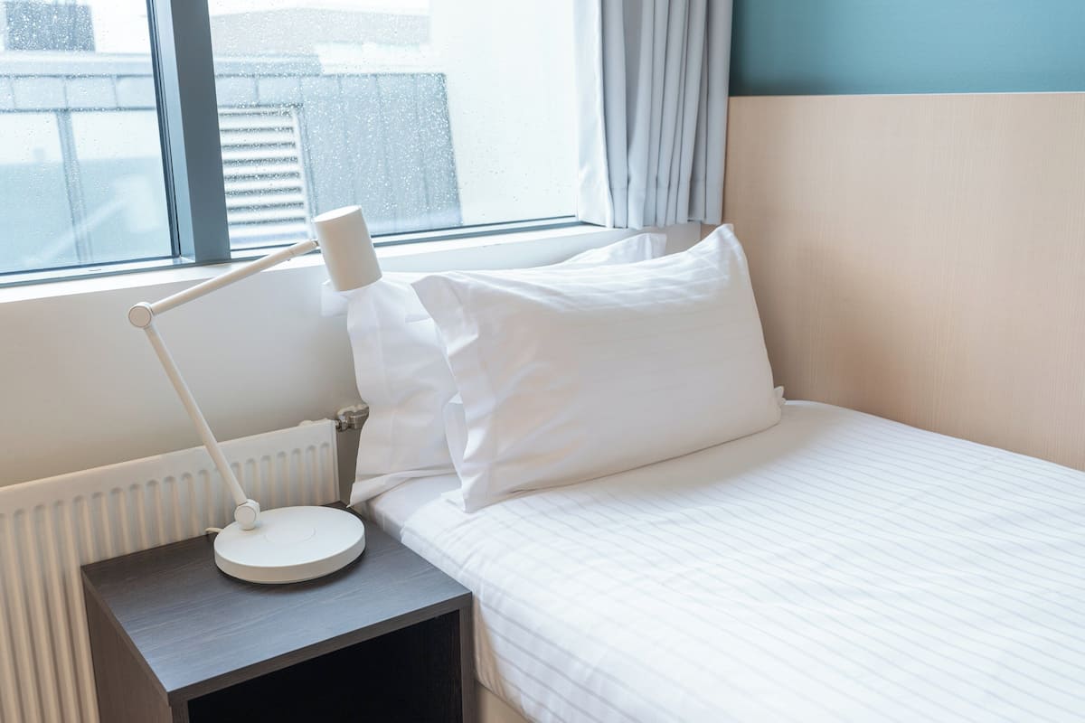 A neatly made bed with white linens, a bedside lamp, and a window with curtains.