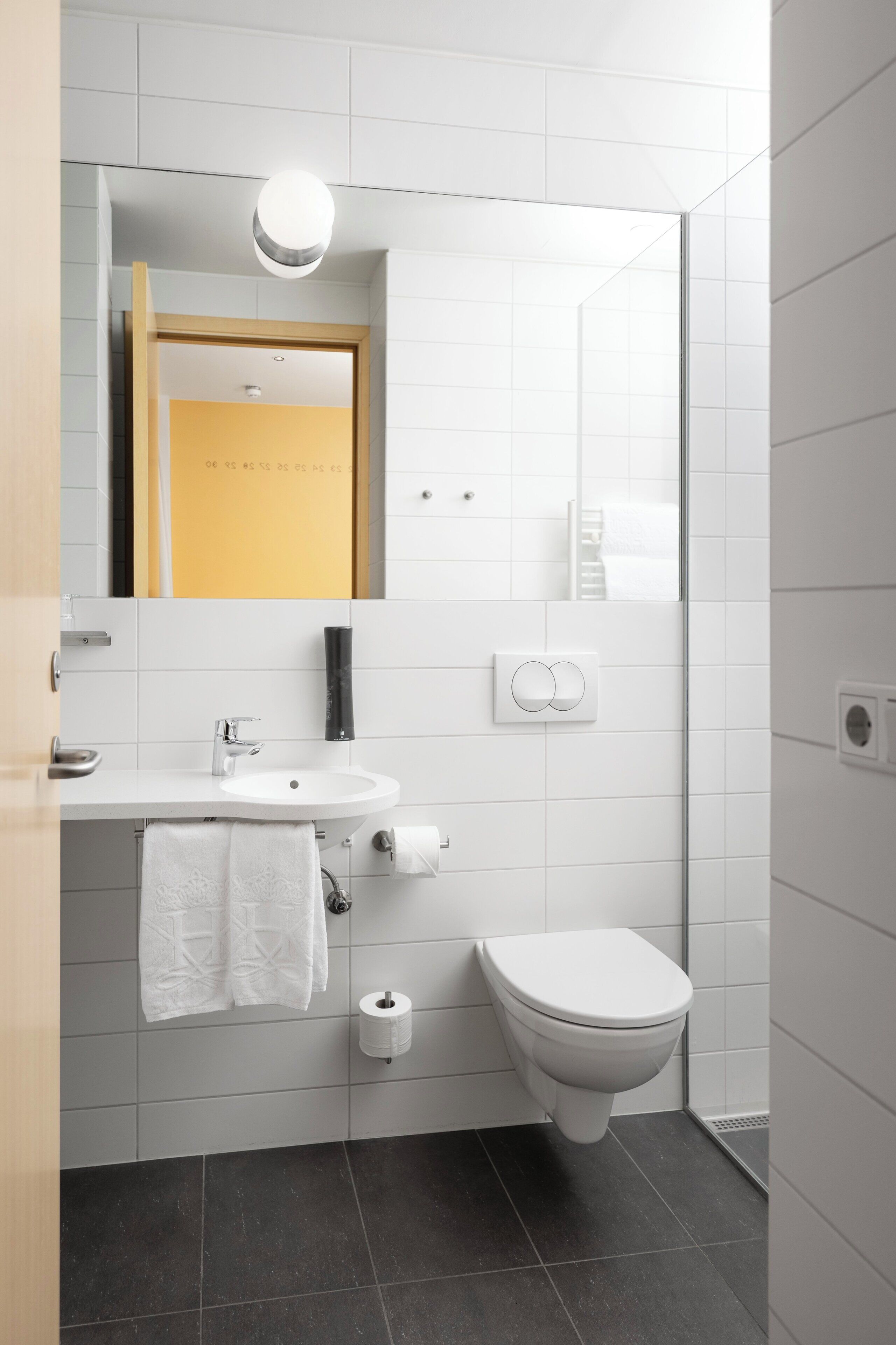 A modern bathroom with a white sink, toilet, and mirror. There is a towel hanging on the wall and a soap dispenser.